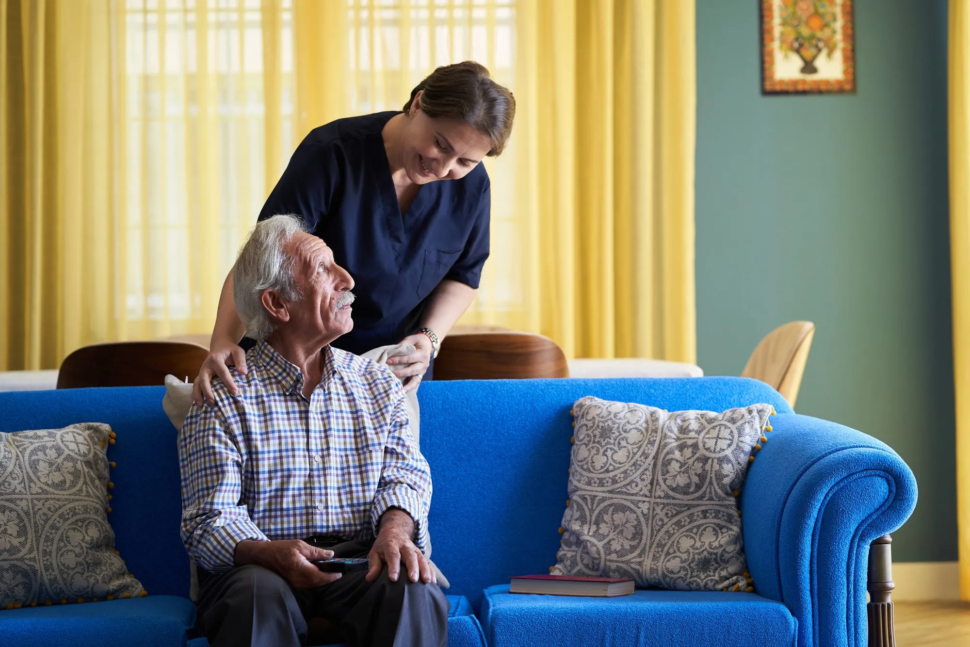 A caregiver checking in with an older man seated on a sofa in his living room.