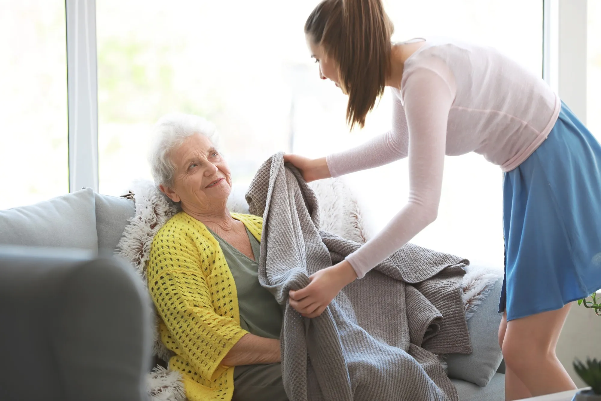 A caregiver placing a blanket over an older woman resting on her sofa.
