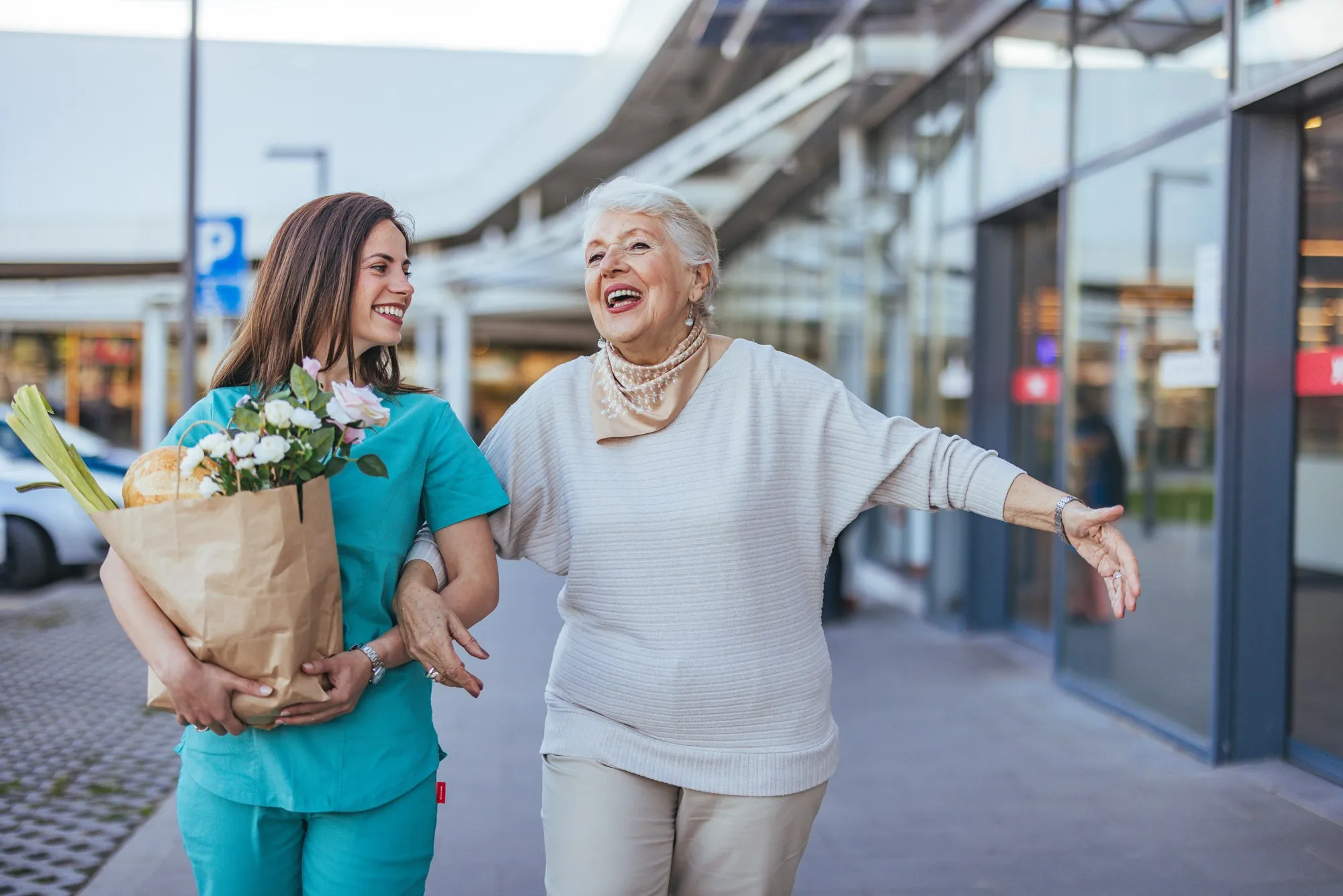 A caregiver carrying a bag of groceries while walking beside a smiling older woman.